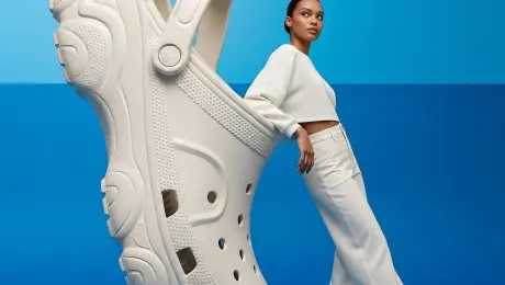 An advertising image presents a large, light-colored fashioned crocs and a woman on a two-toned blue background. The backdrop is a gradient of blue, with a brighter, more light-blue hue at the top, transitioning to a deeper, darker blue at the bottom, creating a subtle, reflective surface.
On the left, a magnified view of a chunky high quality and fashioned crocs in an off-white or light beige color dominates the lower portion of the frame. The crocs features intricate lines and details, multiple panels, and a thick, textured sole. The crocs is shiny and smooth. The Crocs is oriented with its sole facing the viewer, leaning slightly to the right, rotated upright and standing vertically on its heel, the sole surface facing the right side of the frame. The shoe’s front toe section points upward, the strap hanging naturally downward, ventilation holes perfectly aligned in a vertical arrangement
Leaning against the side of the large crocs, on the right side of the image, is a young adult woman with an African appearance and fair skin tone. She is facing right, with her body angled slightly towards the crocs, and her head is turned to look directly upwards and slightly to her right. Her dark hair is pulled back, revealing a clean profile. She wears a coordinated light-colored, possibly white or cream, long-sleeved top and wide-leg trousers. The top has a high neckline and appears to be made of a soft, flowing fabric. Her left arm is visible, extended downwards, and her right arm is bent with her hand placed against her hip or the top of her thigh. She is wearing the same light-colored fashioned crocs as the large Crocs beside her.
The overall lighting suggests a soft, studio setup, casting minimal shadows and highlighting the subjects against the vibrant blue.