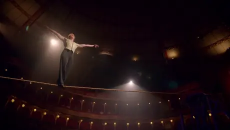 The performer in the image walks steadily on a tightrope high above the circus stage, balancing with arms outstretched, spotlight from below, camera circles upward
