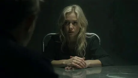 The camera begins in a dimly lit interrogation room, focusing closely on the eyes of a white woman in her early 30s. It slowly pulls back to reveal her full face—platinum blonde hair slightly disheveled, her expression calm but betraying flickers of anxiety. She sits upright in a metal chair, tense wrists resting on the cold tabletop. A harsh overhead light casts sharp shadows across her face, creating stark contrast and strong shadow lines.
