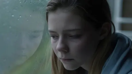 Camera: A stable, slight downward close-up on a young person's face. They are leaning against a window, and their gaze slowly drifts from the window to the middle distance.

Effect: The person's eyes become glossy and wet, but no tears fall. They bite their lower lip for a moment, and their brow furrows slightly. Raindrops slowly trace paths down the window glass, reflecting on their skin.

Sounds/Voices: A soft, melancholic piano melody. The patter of rain against the glass. A single, barely audible sigh.

Mood: Melancholy, contemplative, vulnerable, and introspective.

Lighting: Soft, cool, diffused natural light from the overcast window, giving their skin a pale and somber tone.