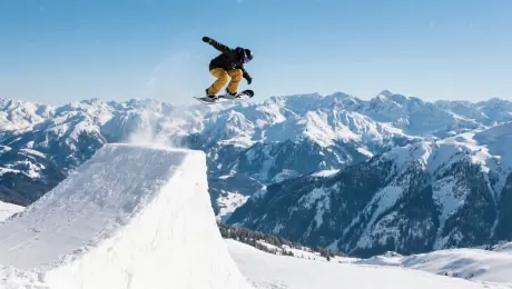 A top snowboarder launches off a massive kicker on a cliff's edge, executing a grab in mid-air. The backdrop is a clear blue sky and a vast mountain range. Powdered snow erupts behind him like smoke in the bright sunlight. Drone-following shot, capturing his perfect form in the air and his smooth landing from a side angle. Extreme sports, 4K, high frame rate, GoPro-like perspective.