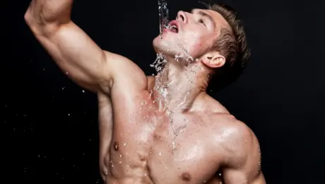A fit man, mid-workout, is captured in a dynamic shot as he drinks water. Sweat glistens on his muscular physique, prominently highlighting his defined abs and biceps. The water he's drinking is dramatically rendered, showing the splash and movement as it enters his mouth, conveying refreshment and exertion.