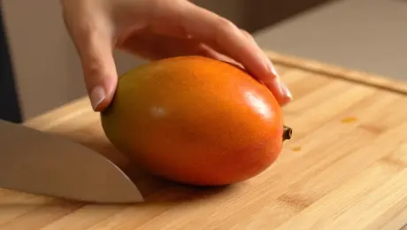 A close-up of hands gently slicing a ripe mango on a bamboo cutting board. The knife glides through the soft flesh with a wet, sticky sound. Juice slowly pools on the surface. The room is silent except for the smooth, squishy slice.