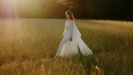 A slow cinematic tracking shot in golden hour light, warm sun rays gently filtering through trees, soft lens flares, delicate depth of field; a young woman in a flowing white dress walks through a meadow of tall grass, petals drifting in the breeze, ultra-realistic textures, gentle camera movement.
