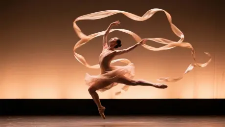 The dancer in the image gracefully moves across the stage with a flowing silk ribbon, spinning and leaping, ribbon twirls through the air, elegant camera dolly-in, warm stage lighting