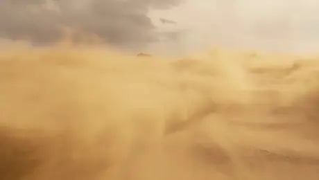 An overhead, cinematic aerial shot sweeps across a vast desert, where endless yellow sandstorms swirl under a raging sky. The wind howls through the barren expanse, filling the frame with an oppressive yet frenzied atmosphere. As a sudden gust surges past the lens, the camera transitions seamlessly, pulling focus onto a lone, veil-covered teenage boy. His figure emerges from the storm, shrouded in mystery, as grains of sand lash across the screen.