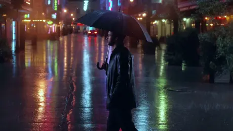 Cinematic panning shot at twilight, rain-soaked streets reflecting neon lights, soft bokeh in the background, warm and cool color contrast; a man stands under an umbrella, slow-motion drizzle, deep shadows and highlights, ultra-detailed photorealism, smooth camera glide.