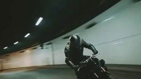 A motorcyclist speeds through the city streets, the third-person camera smoothly tracking from 5 to 10 meters behind. 0–1.5s: He twists the throttle sharply, engine roaring as sparks burst from the wheels. 1.5–3s: Leaping through a gap between skyscrapers, the camera tilts upward from below, capturing the motorcycle arcing through the air with lights streaking across its frame. 3–5s: Landing in a shower of sparks, he crouches low, weaving skillfully through traffic as cars narrowly evade. The bike accelerates into the distance, engine thunder fading away.