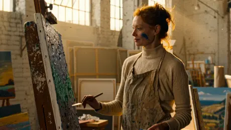 A professional female artist with messy auburn hair tied in a loose bun and smudges of cobalt blue paint on her cheeks, wearing a paint-splattered linen apron over a cream turtleneck, holding a slender palette knife and applying thick impasto texture to a large canvas on a weathered wooden easel, standing in a spacious high-ceilinged loft studio with white brick walls and leaning abstract landscape paintings, voluminous golden sunlight streaming through tall industrial windows with visible dust motes, warm and peaceful creative atmosphere, photorealistic, 8k resolution, shot on 35mm lens, cinematic depth of field, high-fidelity textures of dried oil paint and raw canvas.