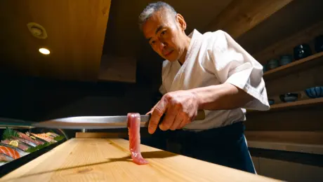 A seasoned sushi chef with salt-and-pepper hair and a concentrated expression, wearing a crisp white cotton uwagi jacket and a dark indigo linen apron, slicing a thin translucent strip of pink fatty tuna with a long polished steel yanagiba knife, standing behind a smooth pale hinoki wood counter in a minimalist traditional Japanese restaurant featuring shelves of handmade ceramic cups and a refrigerated display of fresh seafood, illuminated by warm focused overhead spotlights creating high-contrast shadows and highlighting the glistening texture of the fish, photorealistic 8k resolution, macro photography, sharp focus on the blade edge, cinematic depth of field, shallow bokeh background.
