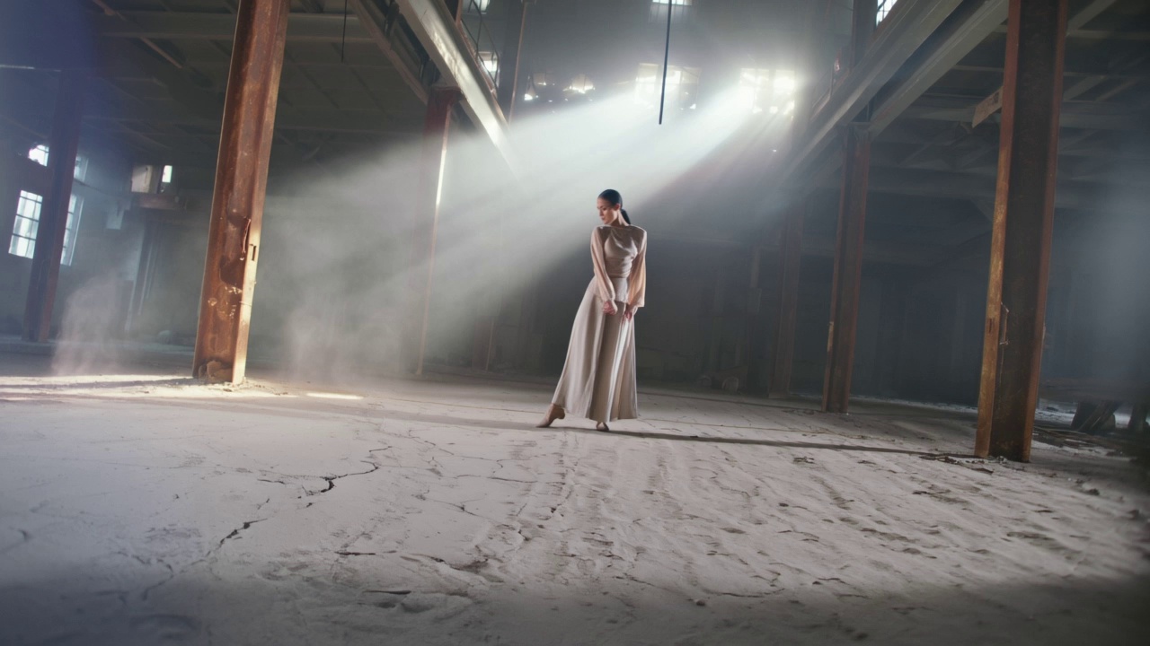 A contemporary dancer in fluid linen clothing performing expressive, rhythmic movements and a slow-motion pirouette, kicking up fine dust from the cracked concrete floor of a cavernous, industrial abandoned warehouse with rusted steel beams. The camera executes a 360-degree dynamic orbiting tracking shot, spiraling upward around the subject to capture the height of the space. Intense volumetric god rays pierce through broken high windows, illuminating swirling dust motes against high-contrast, moody shadows. Cinematic 4K resolution, anamorphic lens flares, shallow depth of field focusing on the dancer’s form, and a gritty, desaturated color grade.