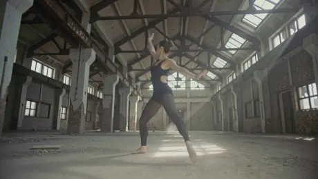 A contemporary dancer in minimalist attire performing fluid, expressive movements including a rapid pirouette and a smooth slide across the dusty floor inside a cavernous, abandoned industrial warehouse with weathered brick walls and rusted structural beams. The camera executes a dynamic 360-degree orbital tracking shot that starts at a low angle and gradually rises to a medium height, following the dancer's flow. Dramatic volumetric light rays stream through broken ceiling skylights, creating high-contrast chiaroscuro shadows and illuminating swirling dust particles in the air. Cinematic 4K resolution, slow-motion 60fps, shallow depth of field with sharp focus on the dancer’s form, gritty industrial aesthetic with subtle organic film grain.