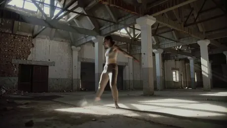 A contemporary dancer in minimalist earth-toned attire performing fluid, high-energy spins and expressive floor work, kicking up subtle layers of dust inside a cavernous abandoned warehouse with exposed brick walls and rusted steel rafters. The camera executes a continuous 360-degree dynamic orbital shot that starts at eye level and transitions into a dramatic low-angle perspective. Intense volumetric lighting streams through broken skylights, creating sharp shafts of light and deep noir-style shadows with visible floating particulates. 4K resolution, high contrast, sharp focus on the subject with cinematic motion blur on the extremities, gritty industrial aesthetic.
