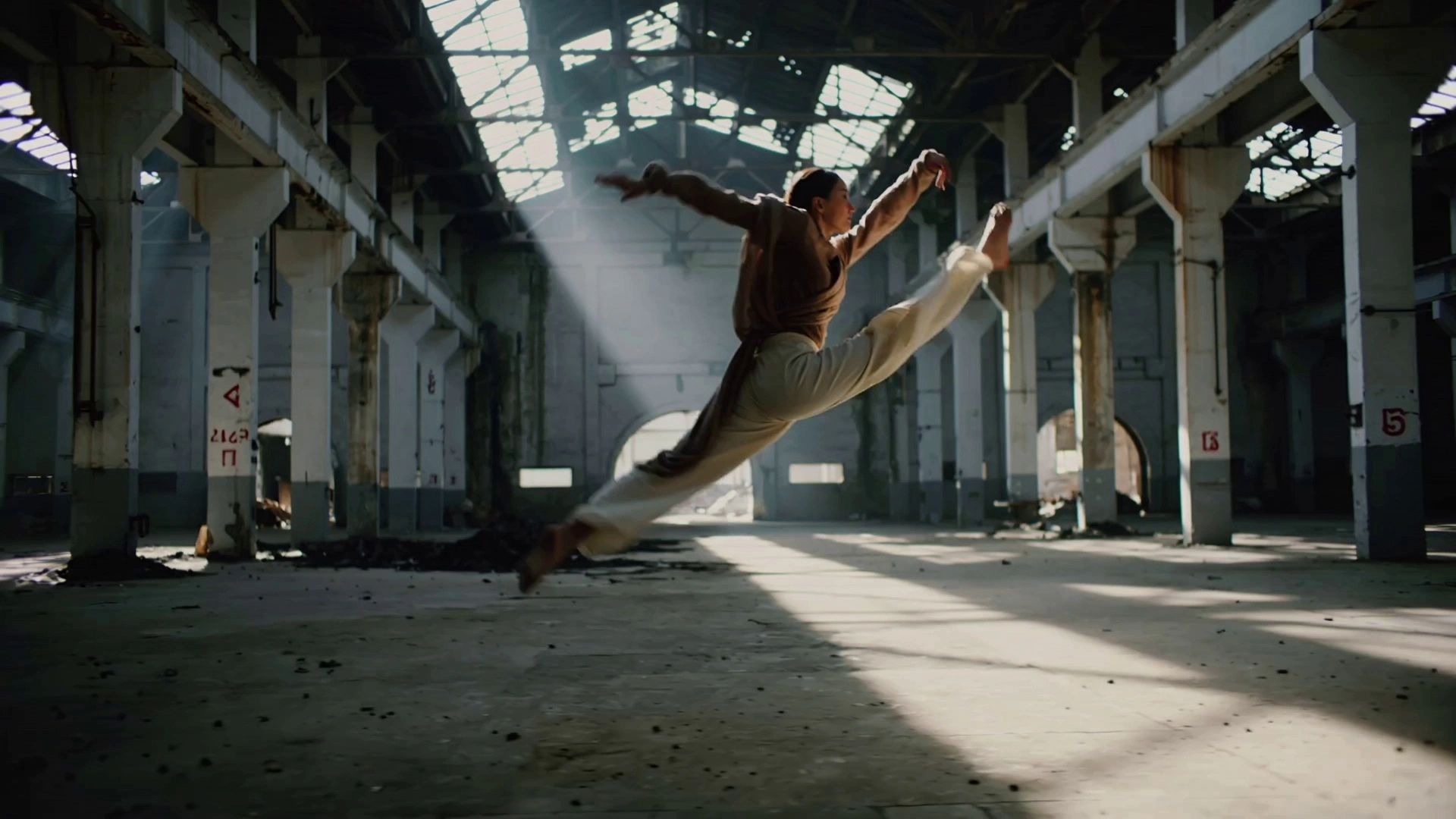 A contemporary dancer wearing flowing, earth-toned linen fabrics, performing a sequence of fluid, athletic leaps and spins with the fabric billowing and trailing their movement. Set within a vast, abandoned industrial warehouse with weathered concrete floors and rusted structural beams. A dynamic orbital tracking shot circles the dancer at a medium distance, capturing the full range of motion while strong volumetric light shafts stream through broken roof panels, creating high-contrast chiaroscuro effects and illuminating swirling dust motes. Cinematic 4K, high-speed capture rendered in fluid slow motion, shallow depth of field with soft bokeh on the industrial background, gritty and atmospheric textures.
