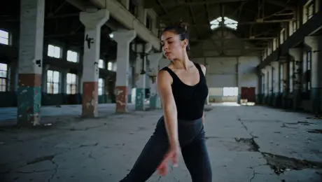 A contemporary dancer in minimalist attire performing a sequence of sharp, fluid movements and a slow, expressive pirouette; a vast, derelict industrial warehouse with cracked concrete floors and rusted steel beams; a continuous 360-degree dynamic orbiting tracking shot that spirals inward toward the subject; volumetric "god rays" piercing through high broken windows, high-contrast chiaroscuro lighting with moody blue shadows and warm golden highlights; 4K cinematic film grain, shallow depth of field with bokeh, 60fps slow-motion for emphasized motion dynamics and swirling dust particles.