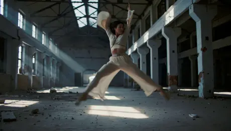 A contemporary dancer in flowing silk attire performing a sequence of high-energy leaps and rapid spins, the fabric trailing through the air inside a vast, derelict industrial warehouse with weathered brick walls and cracked concrete floors. The camera executes a 360-degree orbital tracking shot circling the performer at a dynamic pace. Lighting consists of volumetric light beams piercing through broken ceiling windows, illuminating swirling dust motes with high-contrast chiaroscuro shadows. Visual style includes cinematic 60fps slow motion, anamorphic lens flares, and a shallow depth of field focusing on the dancer's fluid motion.