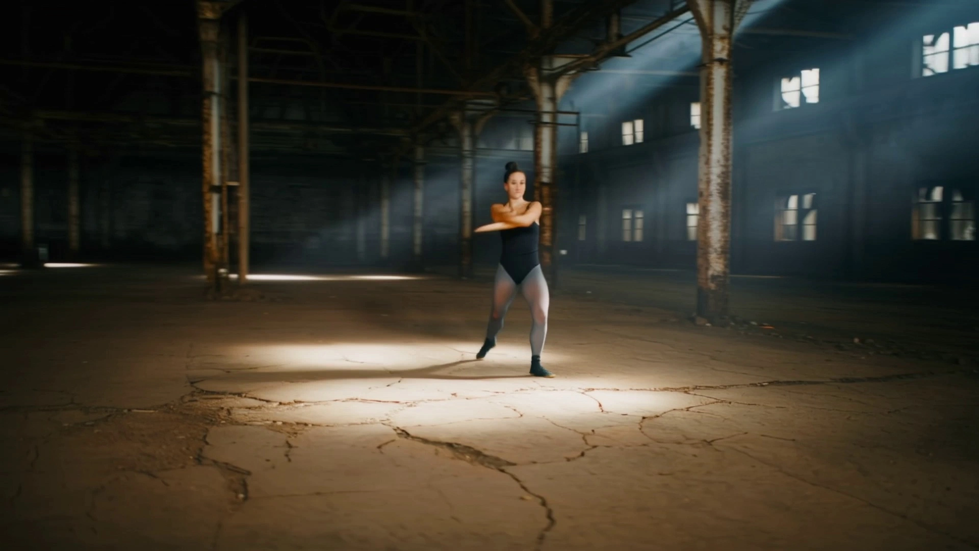 A contemporary dancer performing fluid, high-energy expressive movements and leaps across a cracked concrete floor in a vast, industrial abandoned warehouse with rusted metal beams. The camera executes a dynamic 360-degree orbital tracking shot, circling the performer with smooth, gimbal-stabilized motion. Volumetric god rays stream through high broken windows, creating high-contrast chiaroscuro lighting and illuminating dancing dust motes. Cinematic 4K visual style with a shallow depth of field, sharp focus on the dancer, and rich, gritty industrial textures.