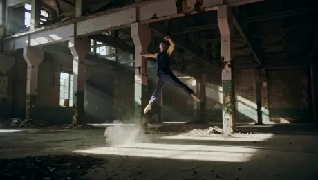 A solo contemporary dancer in fluid motion performing expressive, high-energy leaps and extensions while dust particles swirl around their feet inside a cavernous, derelict industrial warehouse with rusted steel beams and shattered windows; a dynamic 360-degree orbital tracking shot circles the performer to capture the full range of motion; illuminated by sharp volumetric god rays of sunlight piercing through high clerestory windows, creating high-contrast chiaroscuro shadows in a hazy atmosphere; cinematic 4K, slow-motion 60fps, shallow depth of field with sharp focus on the subject, anamorphic lens flares, and a gritty, desaturated color palette.