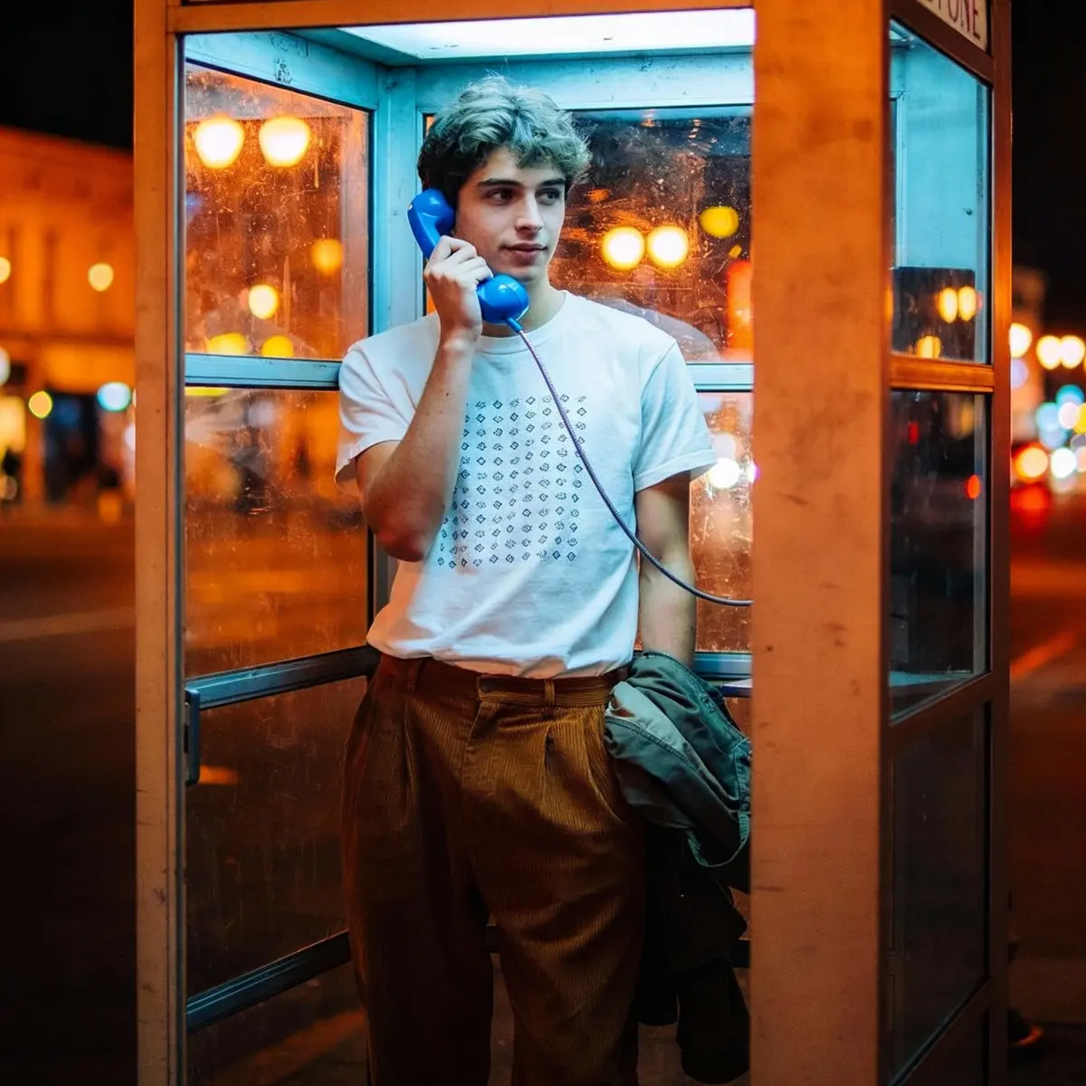 Nighttime outdoor photoshoot: A young man standsinside a public phone booth, holding a blue phonereceiver to his ear. One hand is casually tucked into his pocket, and he strikes a relaxed posture. He wears a white T-shirt with a pattern, loose brown pants, and jacket draped over his arm. The booth's glass reflects city streetlights with a bokeh effect, creating a vintage film style.