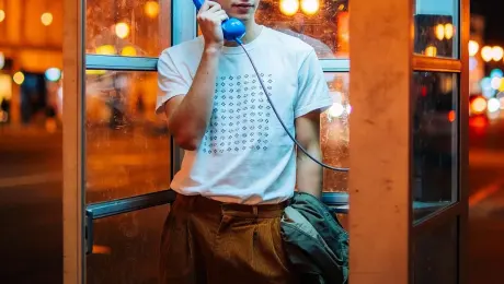 Nighttime outdoor photoshoot: A young man standsinside a public phone booth, holding a blue phonereceiver to his ear. One hand is casually tucked into his pocket, and he strikes a relaxed posture. He wears a white T-shirt with a pattern, loose brown pants, and jacket draped over his arm. The booth's glass reflects city streetlights with a bokeh effect, creating a vintage film style.