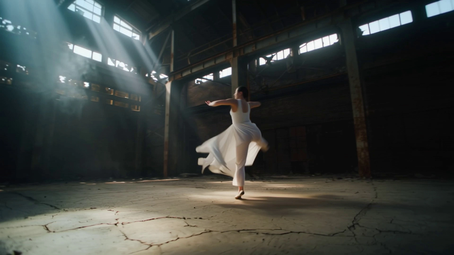 A contemporary dancer in flowing minimalist attire performing fluid, expressive movements including high-velocity spins and controlled floor work. The action unfolds within an expansive, derelict industrial warehouse featuring cracked concrete floors and rusted steel girders. The camera executes a dynamic 360-degree orbit shot, circling the dancer with variable speed to emphasize the peak of each leap, before transitioning into a low-angle tracking shot. Volumetric "God rays" pierce through high, shattered windows, creating high-contrast chiaroscuro lighting and illuminating swirling dust particles that react to the dancer's motion. Cinematic 4K, 60fps slow motion, high dynamic range, shallow depth of field with gritty industrial textures.