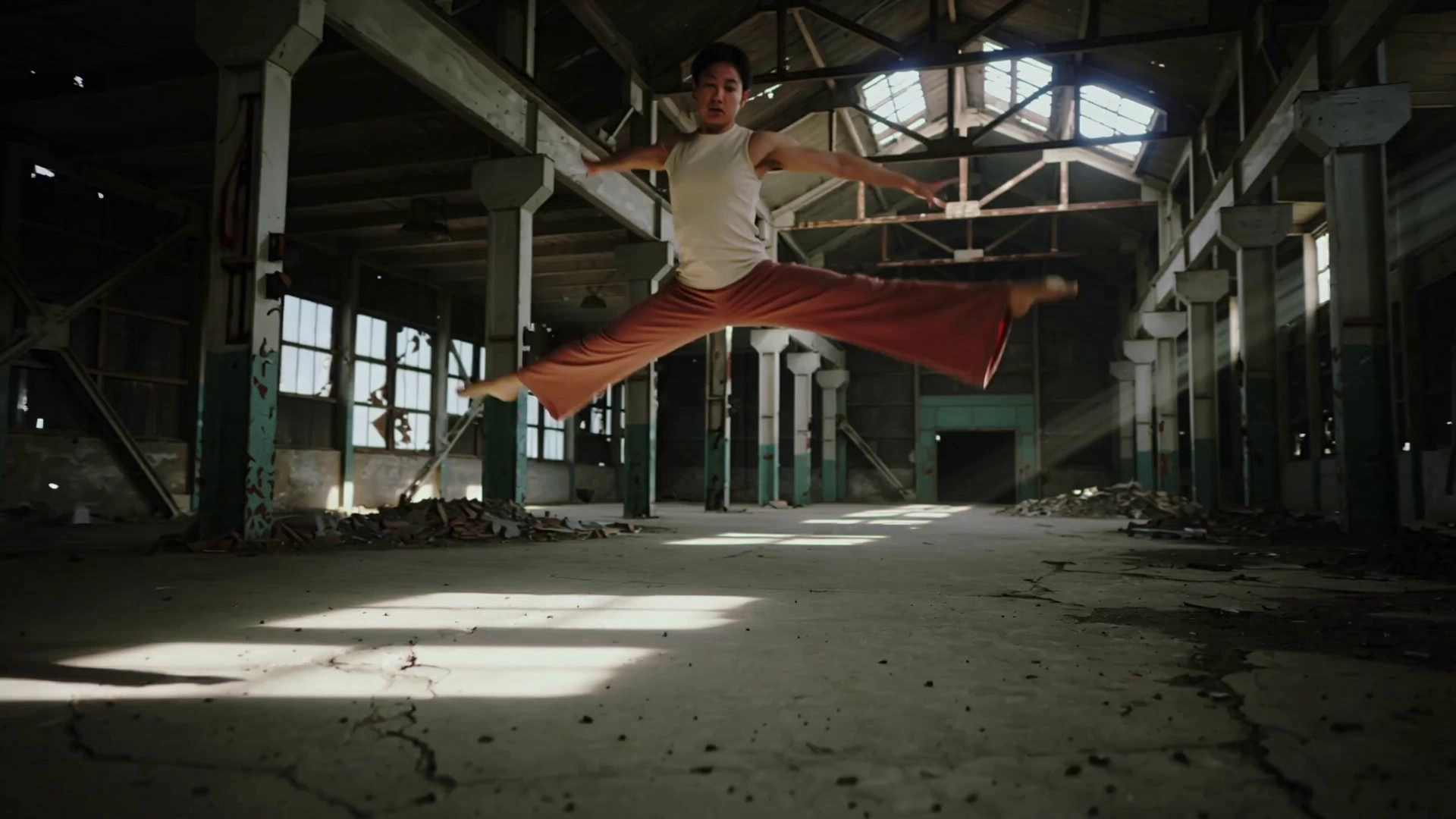 A contemporary dancer in flowing earth-toned attire performing fluid, expressive leaps and intricate floor work within a vast, abandoned industrial warehouse. As the dancer moves, fine dust motes swirl in the air, catching the light. The camera executes a dynamic 360-degree orbital tracking shot, spiraling upward to capture the full range of motion. Dramatic volumetric god rays pierce through high, shattered windows, creating high-contrast silhouettes and long shadows against the cracked concrete floor. Cinematic 4k, shallow depth of field with soft bokeh, slow-motion 60fps capture for emphasized fluid dynamics, anamorphic lens flares, and a gritty, high-detail texture.