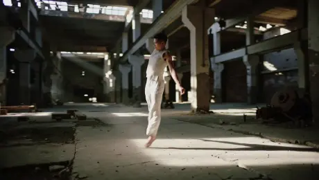 A contemporary dancer in fluid linen clothing performing explosive and graceful movements, leaping and spinning through a cavernous abandoned industrial warehouse with rusted machinery. The camera executes a dynamic 360-degree orbital tracking shot around the subject as they kick up swirling dust particles from the cracked concrete floor. Thick volumetric light beams stream through broken skylights, creating high-contrast chiaroscuro lighting that illuminates the dancer's form against deep shadows. Cinematic 4K, 60fps slow-motion, shallow depth of field with gritty industrial textures and sharp focus on the kinetic energy of the performance.