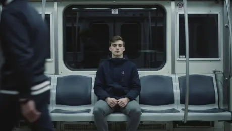 A young man sits motionless in the subway carriage, surrounded by blurred figures hurrying past in a flurry of movement.