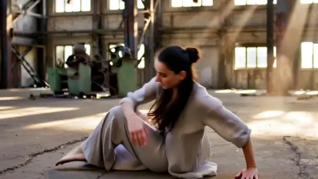 A contemporary dancer in flowing linen attire performing fluid, expressive floor work and an explosive pirouette in a vast, derelict industrial warehouse with cracked concrete and rusted steel beams. A dynamic 360-degree orbital tracking shot circles the subject, capturing the swirling dust motes disturbed by the motion. Dramatic volumetric sunbeams pierce through shattered high-ceiling windows, creating high-contrast chiaroscuro shadows. Cinematic 4K, 60fps slow-motion, shallow depth of field with sharp focus on the dancer’s form, grit-textured film grain, and a desaturated color palette with warm golden highlights.