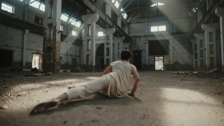 A contemporary dancer in loose linen attire performing fluid floor-work and expressive pirouettes; dust particles erupt from the concrete and swirl rhythmically with the dancer's movements within a vast, derelict industrial warehouse featuring rusted beams and cracked masonry. The camera executes a seamless 360-degree tracking orbit, starting at ground level and ascending into a high-angle crane shot. Dramatic high-contrast lighting with thick volumetric light beams piercing through broken skylights, creating a chiaroscuro effect with deep shadows and golden-hued dust motes. Cinematic 4K resolution, gritty texture, high shutter speed for crisp motion, and a shallow depth of field highlighting the dancer's form against the blurred industrial background.