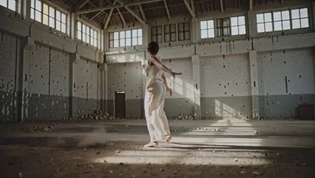 A solo contemporary dancer in minimalist linen attire performing a sequence of fluid pirouettes and expressive leaps, with fabric trailing and concrete dust kicking up from the floor in a sprawling derelict industrial warehouse. The camera executes a high-speed orbital tracking shot circling the dancer 360 degrees, maintaining tight focus on the kinetic motion. Volumetric God rays stream through high clerestory windows, creating a dramatic chiaroscuro effect with high-contrast shadows and brilliant highlights. Cinematic 4K, 60fps slow motion, anamorphic lens flares, and a shallow depth of field emphasizing floating dust motes in the bokeh.