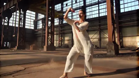 A contemporary dancer in fluid linen attire performing a sequence of sharp, expressive movements and a controlled spin; a cavernous abandoned industrial warehouse with cracked concrete floors and rusted steel beams; a dynamic 360-degree orbital tracking shot that circles the dancer while slowly ascending to a high-angle; volumetric "God rays" piercing through shattered high-altitude windows, illuminating swirling dust particles in high-contrast light and shadow; cinematic 4K, slow motion at 60fps, shallow depth of field focusing on the dancer's form, industrial grit aesthetic.