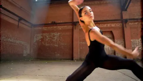 A contemporary dancer in minimalist attire performing fluid, high-energy movements and a controlled slow-motion pirouette inside a derelict industrial warehouse with weathered brick walls. A dynamic 360-degree tracking shot orbits the dancer at mid-range, transitioning from a low-angle to a high-angle perspective. Volumetric "god rays" pierce through broken skylights, creating deep chiaroscuro shadows and illuminating swirling dust motes in the hazy air. Cinematic wide-angle aesthetic, 60fps slow-motion capture, high contrast with sharp detail on fabric textures and gritty concrete surfaces.