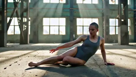 A contemporary dancer in minimalist attire performing a fluid, acrobatic sequence starting from a low floor slide into a powerful mid-air leap, set within a cavernous abandoned industrial warehouse featuring rusted steel beams and cracked concrete. The camera executes a dynamic 360-degree orbital tracking shot that tightens in distance as the dancer reaches the peak of the jump, shifting into a low-angle slow-motion capture. Dramatic god rays pierce through high shattered windows creating high-contrast chiaroscuro lighting with visible swirling dust motes and atmospheric haze. Cinematic 4K resolution, anamorphic lens flares, shallow depth of field with sharp focus on the dancer’s movement, industrial color palette with teal shadows and warm amber highlights.