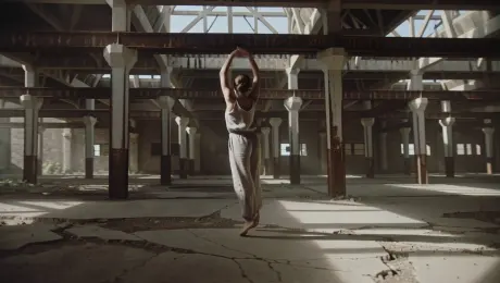 A solo contemporary dancer in flowing linen attire performing fluid, expressive spins and leaps within an industrial abandoned warehouse with cracked concrete floors and rusted beams. A dynamic 360-degree orbital tracking shot circles the dancer as dust particles swirl through high-contrast volumetric lighting and shafts of sunlight piercing through ceiling gaps. The scene features a shallow depth of field, moody atmospheric haze, and 60fps slow-motion playback for enhanced grace and textural detail.