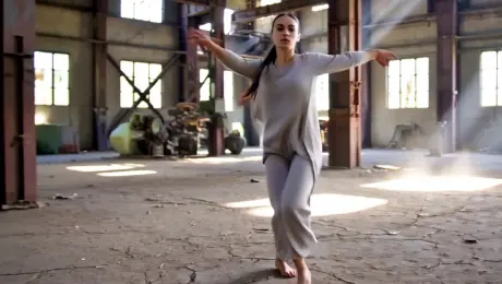 A contemporary dancer in flowing linen garments performing a series of fluid, high-energy spins and expressive leaps across a cracked concrete floor. The scene is set within a vast, derelict industrial warehouse featuring rusted steel beams and shattered windows. The camera executes a dynamic 360-degree orbital tracking shot, circling the dancer at a constant radius as they move through the space. Lighting consists of intense volumetric "God rays" piercing through the ceiling gaps, illuminating swirling dust motes and creating high-contrast silhouettes. Cinematic 4K resolution, captured in 60fps slow motion, with a shallow depth of field that renders the distant machinery into soft, gritty bokeh.