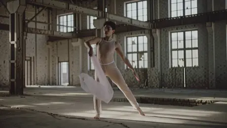 A contemporary dancer in translucent silk garments performing fluid, high-energy expressive movements and sharp contortions; the fabric trails through the air while disturbing floating dust motes in an abandoned industrial warehouse with rusted steel beams and cracked concrete floors. The camera executes a dynamic 360-degree orbiting tracking shot, circling the dancer at a medium distance. Lighting features dramatic volumetric God rays piercing through broken high windows, creating high-contrast chiaroscuro shadows. Visual style is cinematic 4K with a shallow depth of field, high-speed slow motion for motion clarity, and a gritty, atmospheric texture.