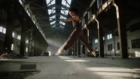 A contemporary dancer in fluid, earth-toned attire performing an expressive leaping and floor sequence, limbs extending with explosive grace as dust motes swirl around their movement, set within a vast abandoned industrial warehouse featuring cracked concrete and rusted steel girders; the camera executes a continuous 360-degree orbital tracking shot that circles the dancer, maintaining a medium-wide frame while subtly descending; volumetric God rays pierce through high clerestory windows to create high-contrast chiaroscuro lighting and sharp shadows; cinematic 60fps slow motion, anamorphic lens flares, gritty industrial texture with a shallow depth of field and sharp focal clarity on the dancer's form.