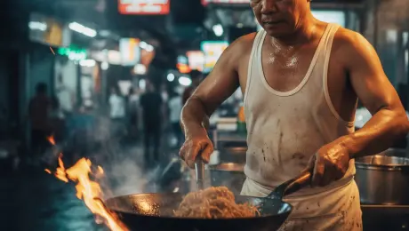 Middle-aged street food chef in a white tank top vigorously stir-frying noodles in a seasoned wok, intense orange flames leaping from the burner and flickering dynamically, thick white steam and smoke billowing and swirling around his sweating face, glistening sweat droplets trickling down his skin. Cinematic slow zoom-in with subtle handheld camera shake, shallow depth of field with blurred neon city signs and distant crowd movement in the background. Gritty night market atmosphere, volumetric lighting, 4k, 60fps, slow motion, cinematic color grading, photorealistic textures.