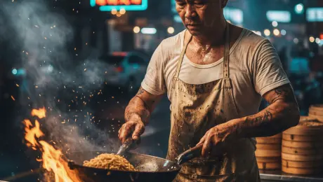 The weathered street food chef with visible tattoos vigorously tosses a wok full of noodles over an intense, leaping flame. Sweat glistens and drips from his forehead as he works with rhythmic intensity, while thick clouds of steam and smoke swirl upward, mixed with glowing orange sparks flying from the burner. The camera executes a low-angle slow push-in with a subtle handheld camera shake to capture the raw energy of the heat. Cinematic contrast between the warm, flickering orange firelight on the chef's skin and the cool blue and cyan neon street signs in the blurred night market background. 4k, 60fps, slow motion, motion blur, highly detailed, realistic texture, cinematic color grading, volumetric lighting, photorealistic.