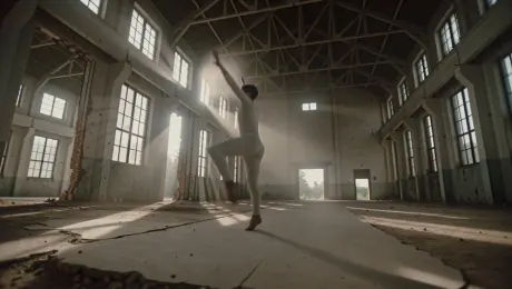 Contemporary dancer in fluid garments performing explosive leaps and rhythmic floor work amidst plumes of suspended dust within a derelict industrial warehouse. The camera executes a dynamic 360-degree orbital tracking shot at a low angle, following the dancer's centrifugal motion. Lighting consists of high-contrast chiaroscuro with dramatic volumetric light rays piercing through shattered high-vaulted windows, casting elongated shadows on the cracked concrete floor. Cinematic 4K, high shutter speed for motion clarity, and a gritty, desaturated color palette with warm golden dust particles.