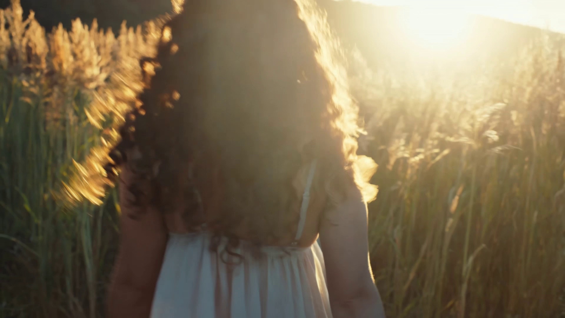 Golden hour shot. A young woman with curly hair walking in a field of tall grass turns back to look at the camera and smiles genuinely. The low sun is behind her, creating a beautiful rim light (backlight) on her hair. Dust motes dancing in the light. Handheld camera feel, warm and nostalgic.