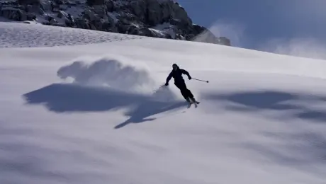 A skier glides downhill, carving turns that kick up clouds of snow. The camera follows steadily as he picks up speed along the slope.