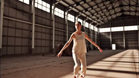 A contemporary dancer in flowing linen attire performing fluid, expressive movements and sharp leaps in a cavernous, industrial abandoned warehouse. Fine dust particles rise from the cracked concrete floor with every footfall, swirling into the air. The camera executes a dynamic 360-degree orbital tracking shot, circling the dancer to capture the full range of motion. High-contrast volumetric "God rays" stream through broken overhead windows, creating dramatic light paths and illuminating the airborne motes. Cinematic slow motion, shallow depth of field with sharp focus on the subject's expressions, gritty industrial textures, and 4K hyper-realistic detail.