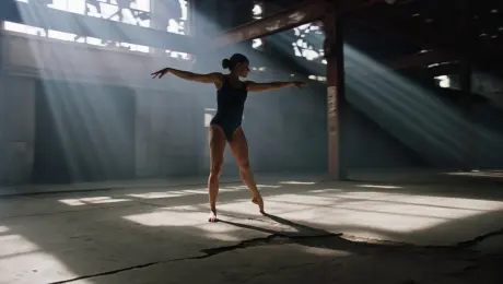 A contemporary dancer performing a sequence of fluid, expressive movements and leaps through an abandoned industrial warehouse. The camera executes a dynamic 360-degree orbital tracking shot, circling the performer while maintaining a tight focus. The environment is characterized by rusted steel beams, cracked concrete, and shattered windows. Dramatic volumetric light beams stream through the ceiling, creating high-contrast chiaroscuro shadows and highlighting the dancer's silhouette. Cinematic 4K resolution, slow-motion 60fps frame rate, shallow depth of field with sharp subject isolation, and a gritty, desaturated color palette.