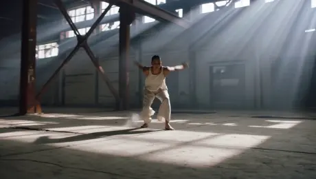 A contemporary dancer in fluid linen attire performing an expressive sequence of high-energy leaps and intricate floor-work, with swirling dust motes rising from the impact, set within a vast abandoned industrial warehouse featuring cracked concrete and rusted steel beams. The camera executes a dynamic 360-degree orbital tracking shot at a low angle, circling the subject to capture the full range of motion. Volumetric god rays stream through high, broken windows, creating high-contrast chiaroscuro shadows and a thick atmospheric haze. Cinematic 4K aesthetic, high frame rate for subtle slow-motion clarity, sharp focus on the dancer with a shallow depth of field blurring the gritty industrial background.