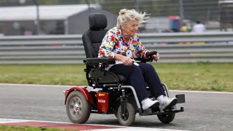 Both accelerate hard out of a corner together — grandmother's wheelchair wobbles at the limit, she counter-steers with pure instinct, knuckles white on the joystick, her reading glasses flying off her face into the slipstream. The F1 driver glances over through his visor, visibly shaken. She doesn't look back. They hit the straight — the F1 car pulls away slowly, but she tucks into its wake and drafts, inching back up. Cut to the grandstands erupting.