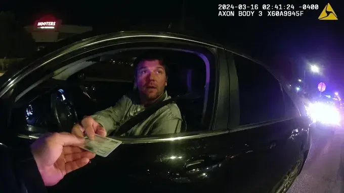 From the perspective of a police body-worn camera in the early hours of the morning, a driver hands over their license. The footage bears a watermark and a timestamp.