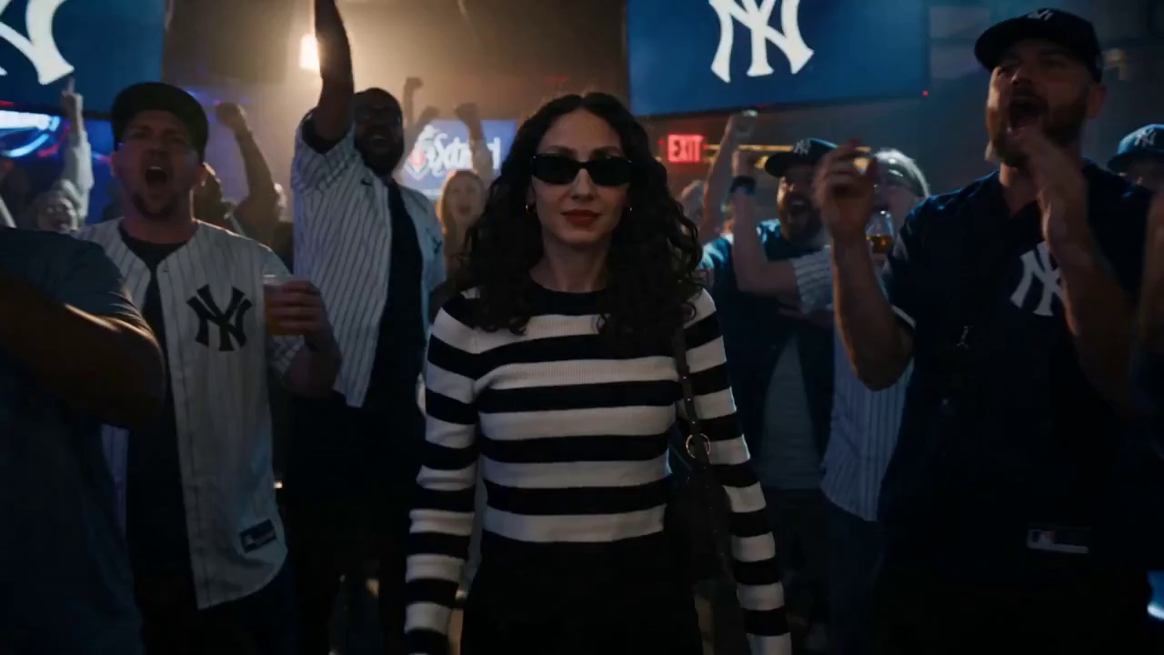 Use @ Reference Image as the main character, keeping facial features and body proportions consistent throughout. She is a 30-year-old women. Cinematic time-freeze short film, 15 seconds, ultra-realistic, shot on Arri Alexa Mini, 35mm lens, moody sports bar interior lighting with neon accents, volumetric haze, dynamic hard shadows, shallow depth of field.

[0:00–0:03] A packed, dimly lit sports bar, time flowing normally. Steadicam frontal medium shot tracking her walking confidently through absolute chaos. On the blurred TVs in the background, the Yankees have just clinched the World Series. The crowd is erupting in pure euphoria. As he walks, he raises his right hand and snaps.

[0:03–0:06] At the snap, a subtle spherical shockwave bursts from her fingertips with air distortion and light refraction, expanding outward. Everything freezes mid-explosion. Thick arcs of golden beer suspend in the air, with individual glistening droplets visible. Popcorn kernels float motionless. People are frozen mid-cheer and mid-air in leaping embraces. Volumetric neon light catches the suspended dust and liquid. Absolute silence.

[0:06–0:09] Only she moves. her footsteps echo slightly. The camera moves backward as he walks through the frozen celebration, observing calmly. she casually ducks under an arc of frozen, splashing beer. she notices a cluster of floating popcorn, reaches out, and plucks a single kernel from the air.

[0:09–0:11] sHe stops in front of a frozen women wearing a Yankees jersey, her face locked in an ecstatic, tearful scream, arms raised for a high-five. The main character tilts her head, gently adjusts the brim of the frozen women's Yankees cap, nods slightly, and softly says: “perfect.”

[0:11–0:15] She turns, tosses the popcorn kernel into her mouth, and crunches it. She faces the camera, smirks, and snaps again. A stronger reverse shockwave restores motion. The beer completes its splash, the cheering roar instantly returns, and fans land their jumps. She walks away as the camera pushes through the wildly celebrating crowd. Fade to black.

Sound: deafening bar roar/celebration → snap → deep shockwave/bass drop → absolute silence → footsteps → sharp popcorn crunch → “perfect” → snap → reverse shockwave → deafening celebration returns.