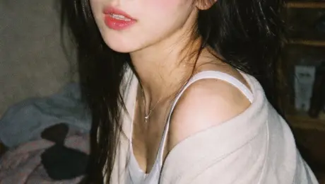 Generates a highly realistic, candid portrait with a vintage film aesthetic and direct flash lighting.

A vintage 35mm film photograph of a {argument name="subject description" default="young Asian woman"} with {argument name="hair style" default="long dark wavy hair and wispy bangs"}. She is wearing a {argument name="clothing" default="white ribbed tank top and a loose beige knit cardigan slipping off one shoulder"}, along with a delicate silver necklace. She has soft makeup with pink blush and glossy lips, looking directly at the camera with slightly parted lips. The lighting is harsh direct camera flash, creating a candid, amateur snapshot aesthetic. The background is a {argument name="setting" default="dimly lit, slightly messy room with clothes on a table and a wooden shelf"}. The image features heavy film grain, slightly muted colors, and a nostalgic, highly realistic photographic texture.